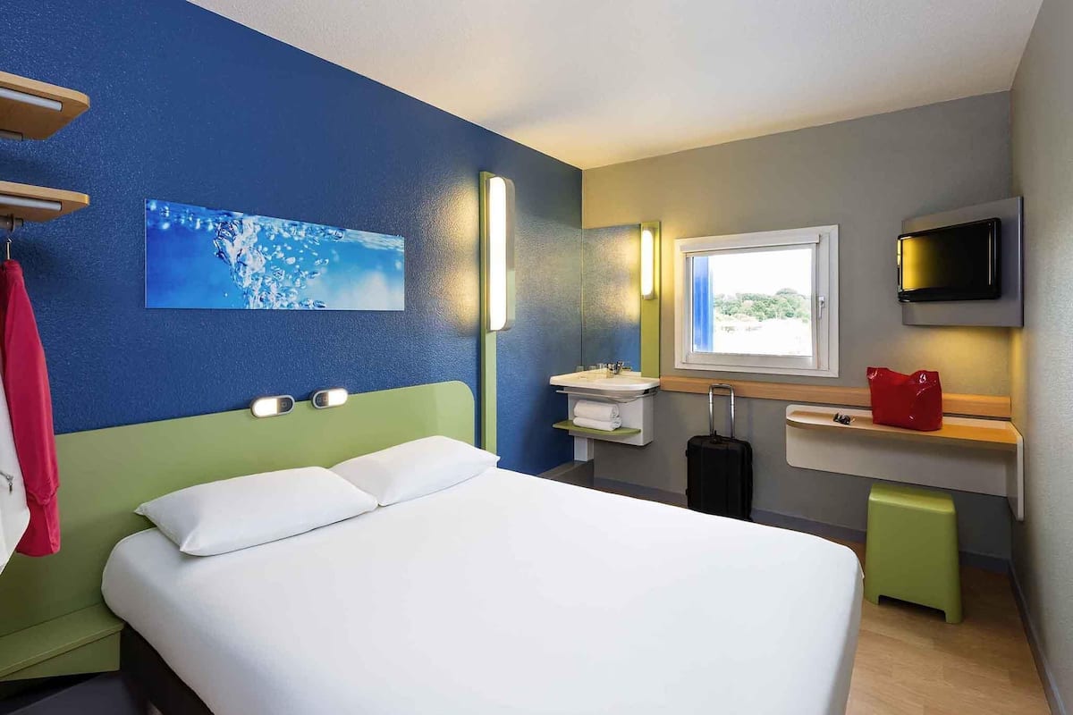 A hotel room with a bed, a bathroom with a sink and mirror, a TV, and a window.