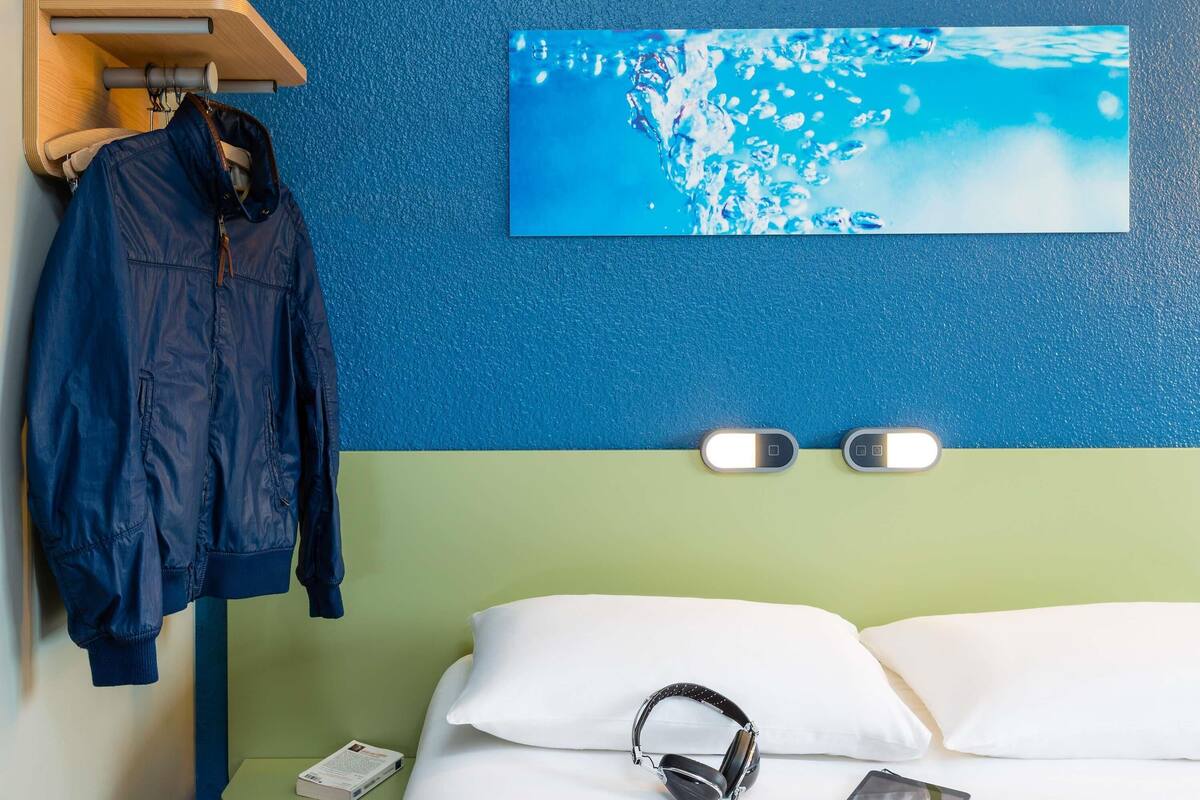 Une chambre d’hôtel avec un lit, une veste bleue suspendue à un porte-manteau, un tableau représentant de l’eau accroché au mur et deux lampes fixées au mur.