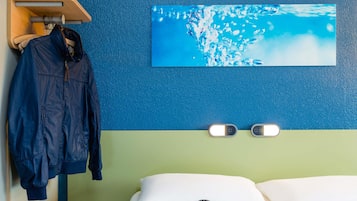 Quarto de hotel com uma cama, um casaco azul pendurado em um suporte, uma pintura de água na parede e duas luzes fixadas na parede.