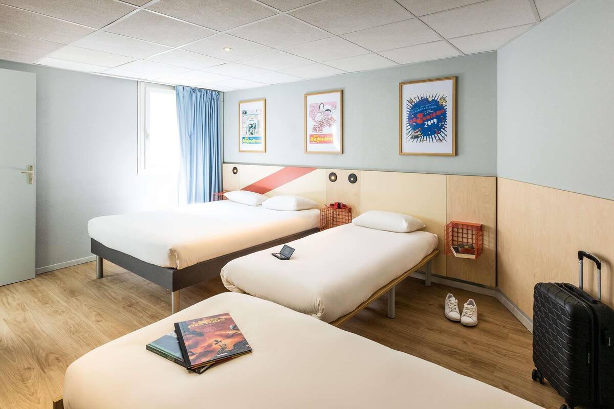 Quarto de hotel com duas camas, uma escrivaninha, uma cadeira, uma mala e uma janela com cortinas.
