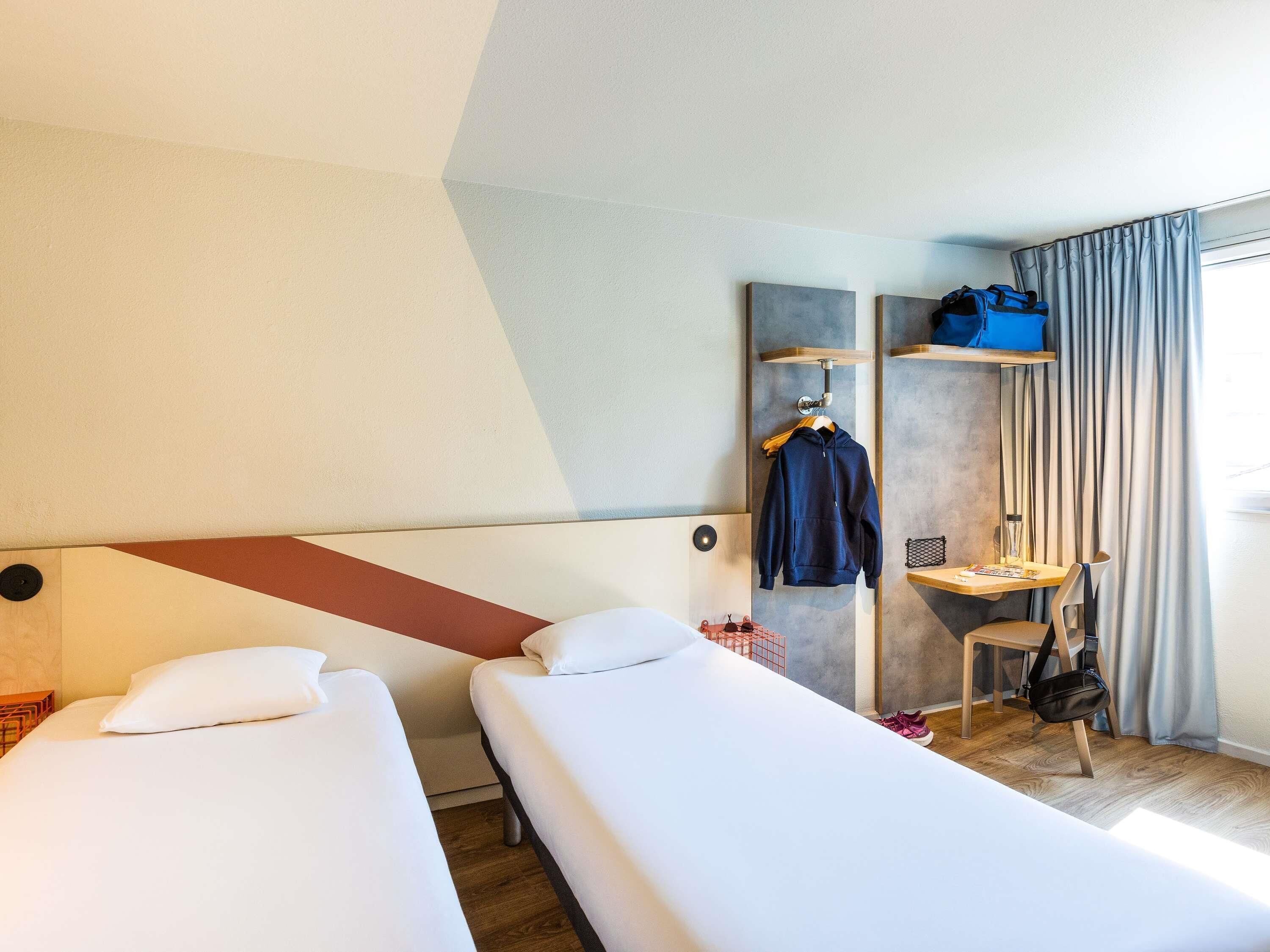 A hotel room with two beds, a desk with a chair, a hanging coat rack, and a window with curtains.