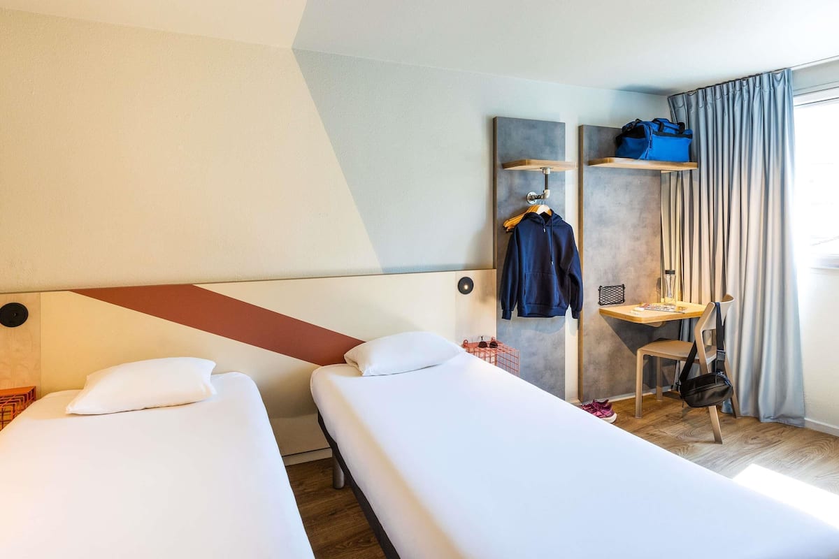 Ein Hotelzimmer mit zwei Betten, einem Schreibtisch mit Stuhl, einem Kleiderhaken und einem Fenster mit Vorhängen.