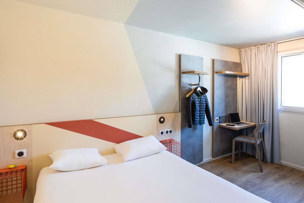 Quarto de hotel com uma cama grande, uma escrivaninha com um laptop, uma cadeira e um porta-roupas com um casaco.