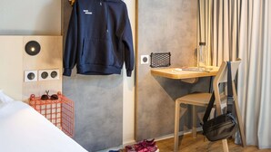 Habitación de hotel con una cama, un escritorio con silla, perchero, zapatero y toma de corriente en la pared.