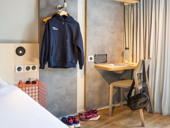 A hotel room with a bed, a desk with a chair, a hanging coat rack, a shoe rack, and a wall-mounted power outlet. - ibis budget Angoulême Centre (Angouleme)