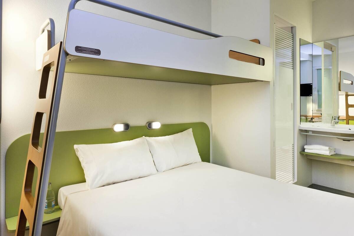 Un lit superposé avec une tête de lit verte, une table de chevet avec une bouteille, et un miroir avec une étagère au-dessus.
