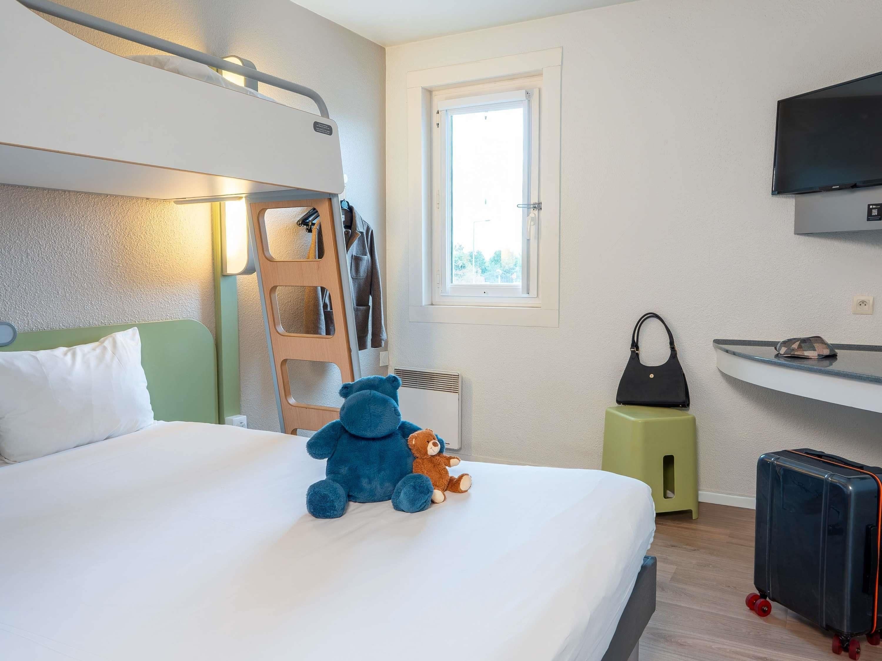 A hotel room with a bunk bed, a small desk, a television, a window, and a suitcase.