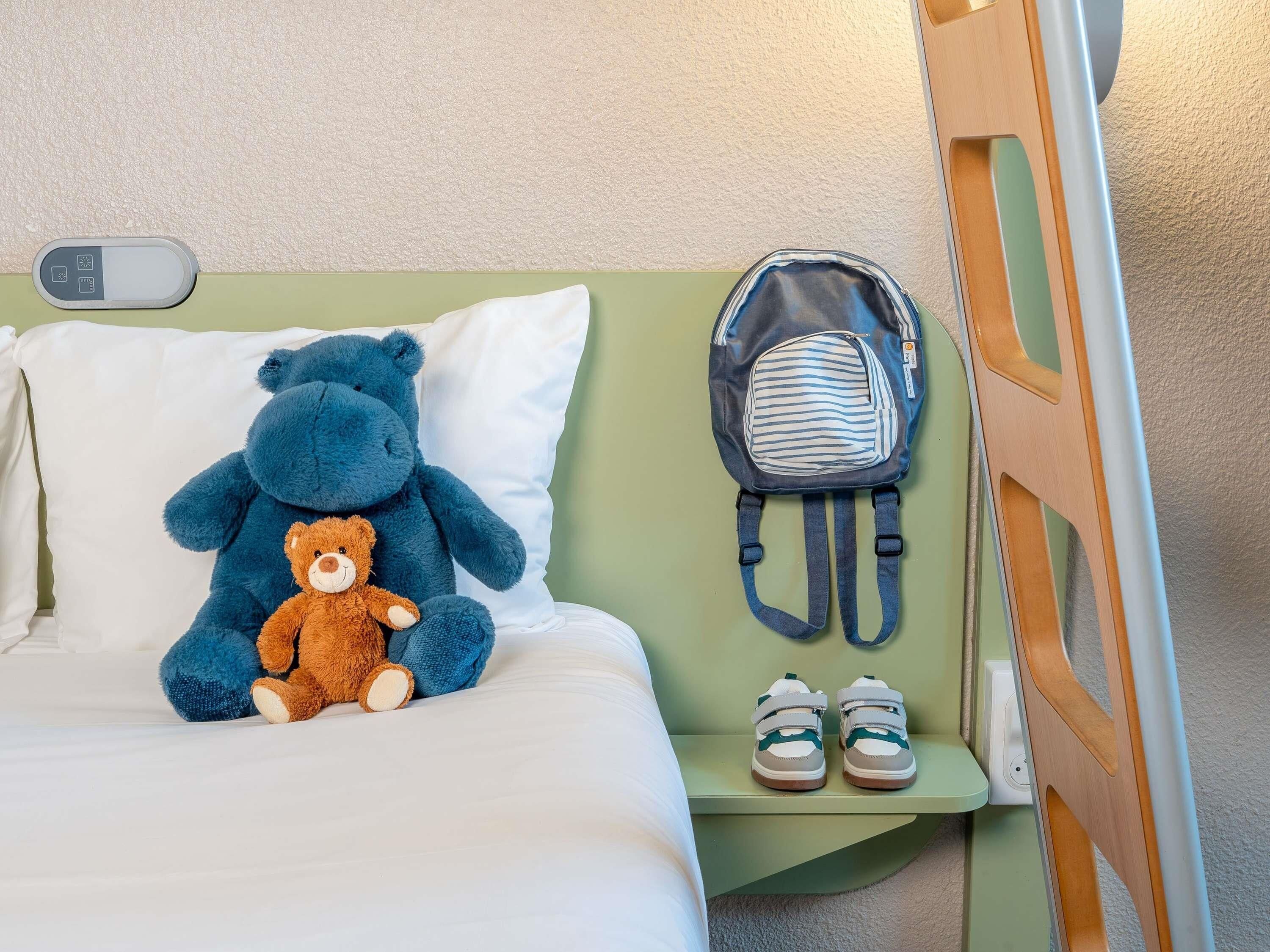 Una cama con un oso de peluche, una mochila a rayas y un par de zapatos sobre una mesita de noche.