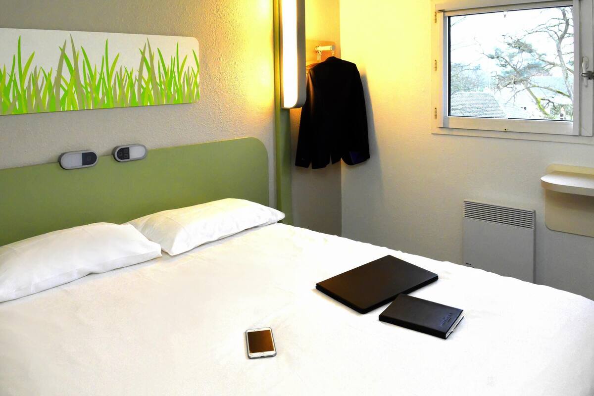 A hotel room with a bed, pillows, a bedside table with a lamp, a window, and a wall-mounted artwork.