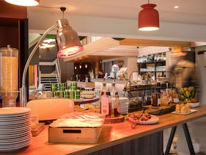 A buffet setup with various food items, a beverage station, and a bar area in the background. - ibis budget Rouen Sud Zenith (Saint-Etienne-du-Rouvray)