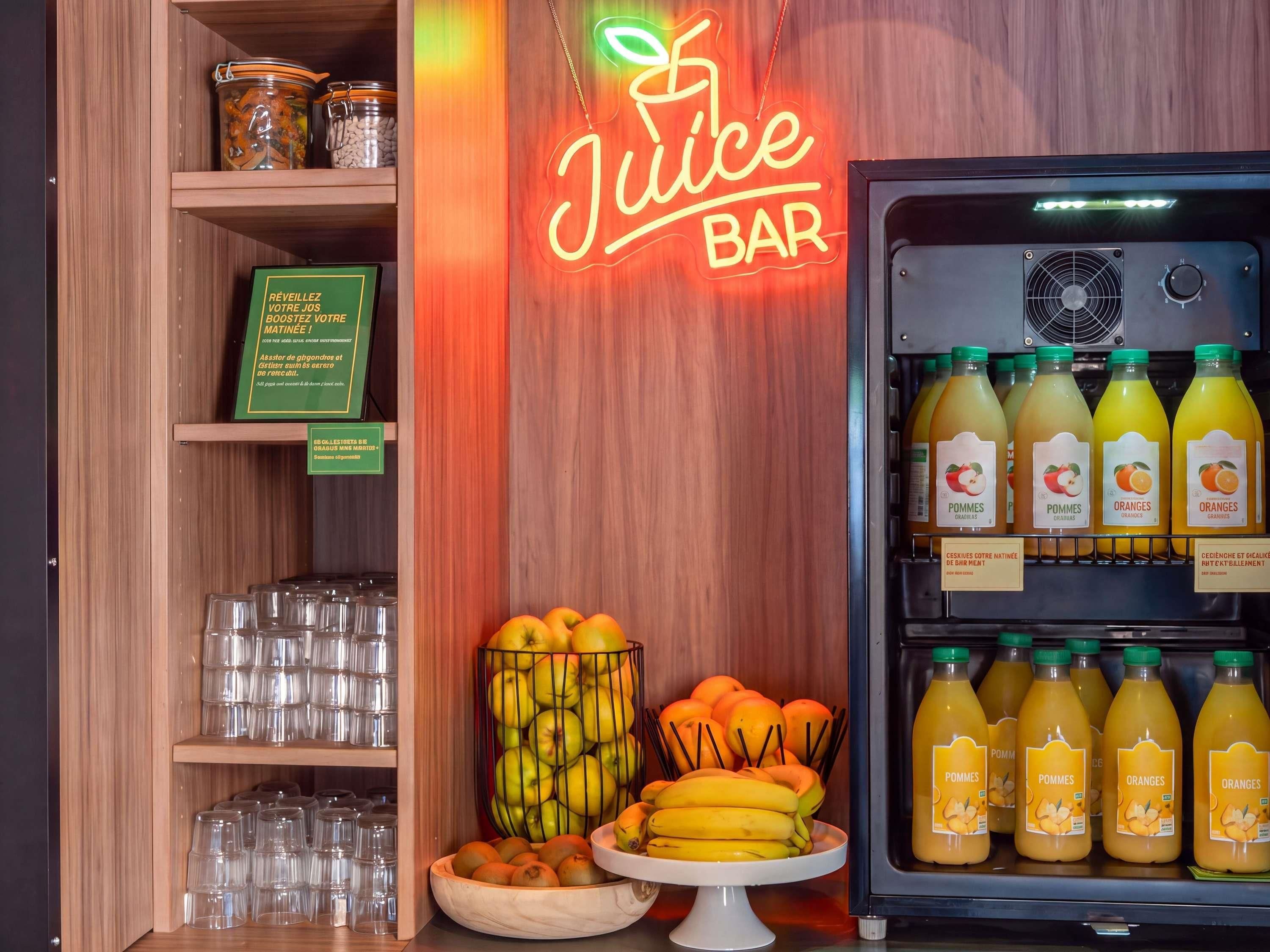 Un bar à jus avec une enseigne au néon, un présentoir réfrigéré rempli de jus de fruits et une variété de fruits et de verres disposés sur des étagères.