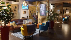 Een moderne lobby met een leren bank, kleurrijke stoelen, een grote potplant en een tv aan de muur.