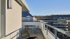 Een balkon met buitenmeubilair, een hekwerk en uitzicht op gebouwen in de verte.