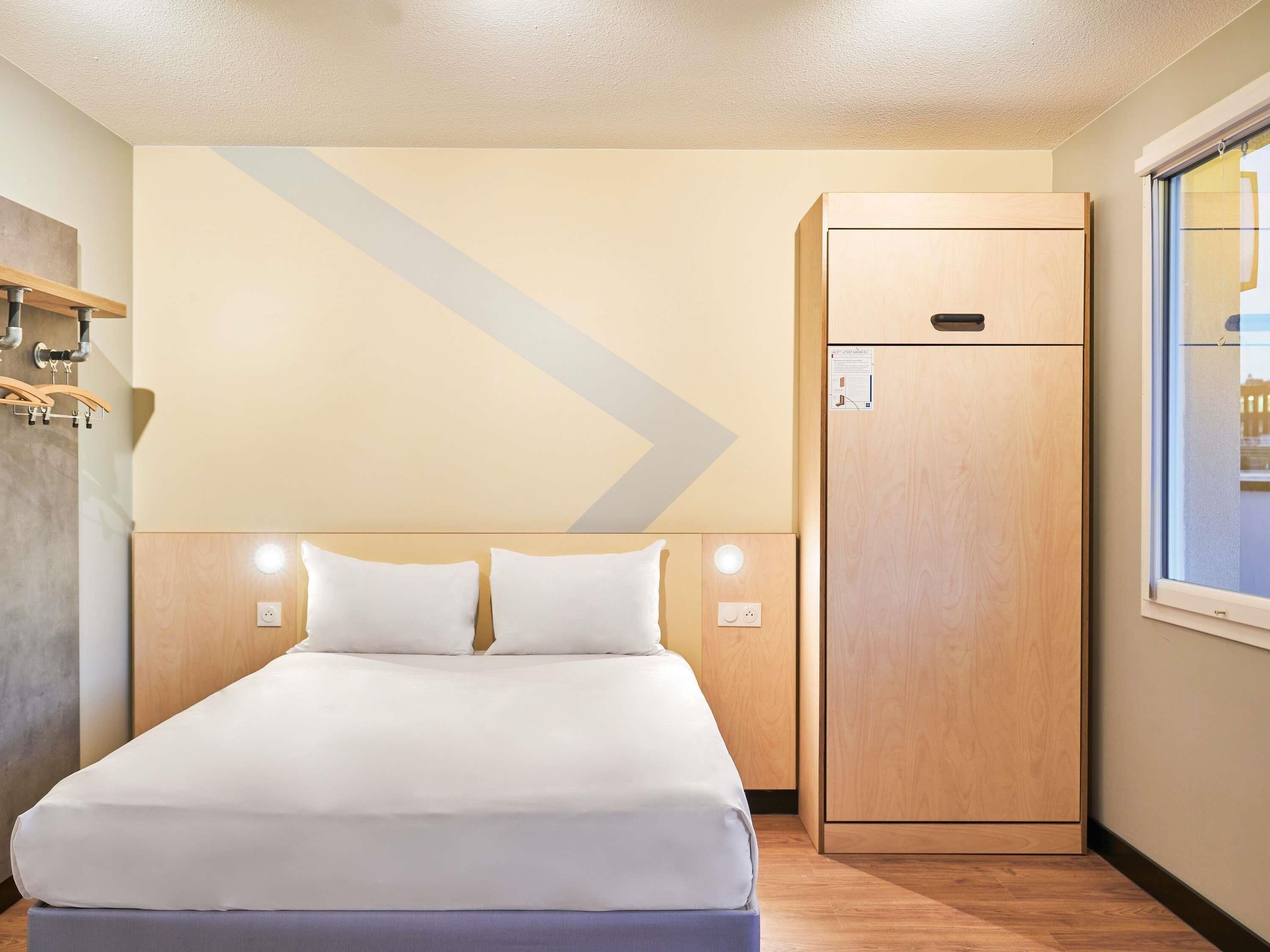 A hotel room with a bed, a wardrobe, a window, and a wall-mounted light fixture.