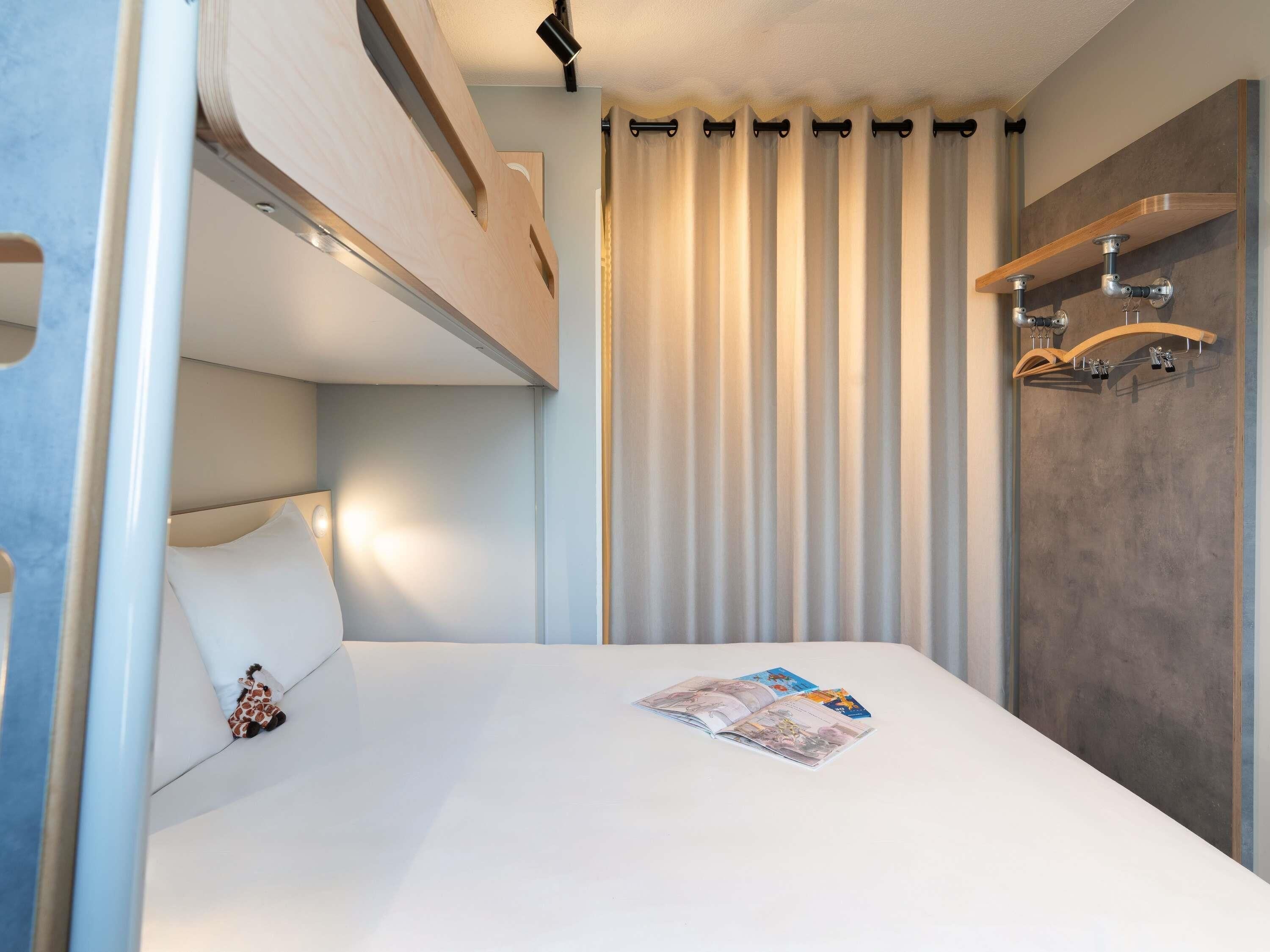 Uma cama beliche com cabeceira, uma mesa de cabeceira com luminária, uma revista na cama e uma cortina com um lustre acima.