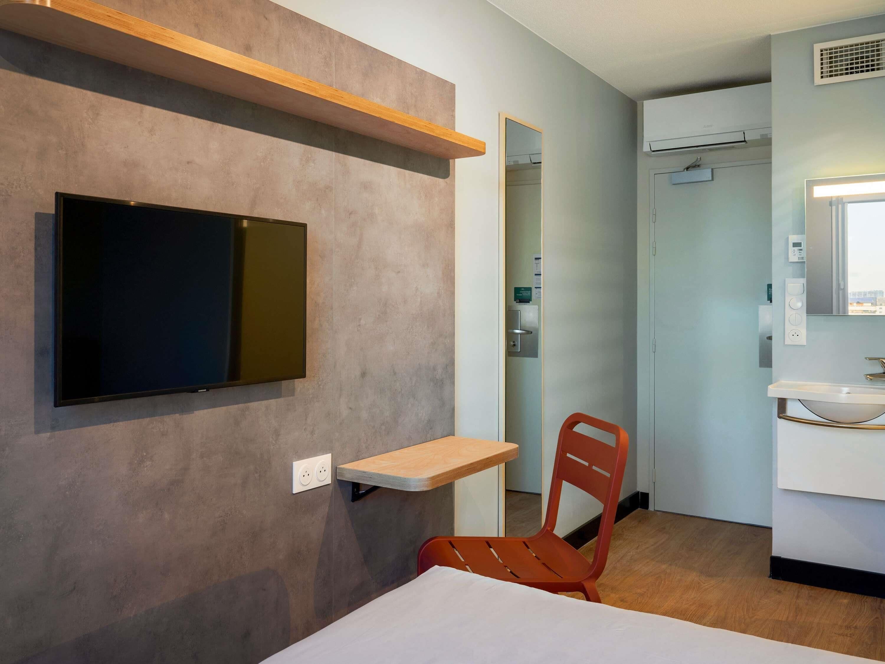 Ein Hotelzimmer mit einem Bett, einem Wand-Fernseher, einer kleinen Holzablage, einem roten Sessel, einem Badezimmer mit Waschbecken und Spiegel sowie einer Tür zu einem Schrank.