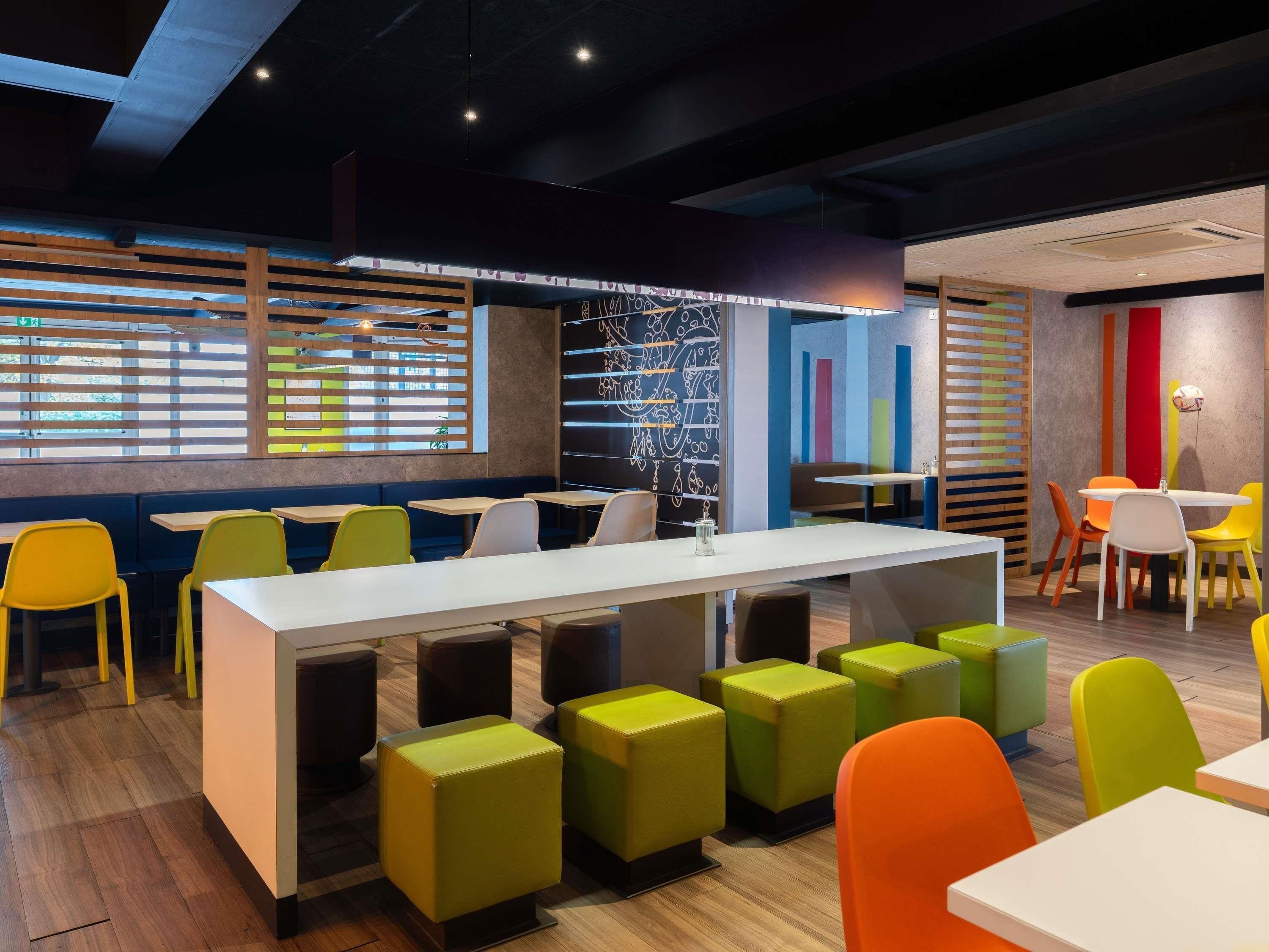 A modern restaurant interior with a long communal table, colorful chairs, and a vibrant wall mural.
