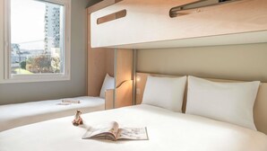 Hotelkamer met een stapelbed, een raam met uitzicht op gebouwen, een nachtkastje met een boek en een giraf speelgoed, en een hoofdeinde met een lamp.
