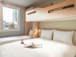 Ett hotellrum med en våningssäng, ett fönster med utsikt över byggnader, ett nattduksbord med en bok och en leksaksgiraff, och en sänggavel med en lampa.