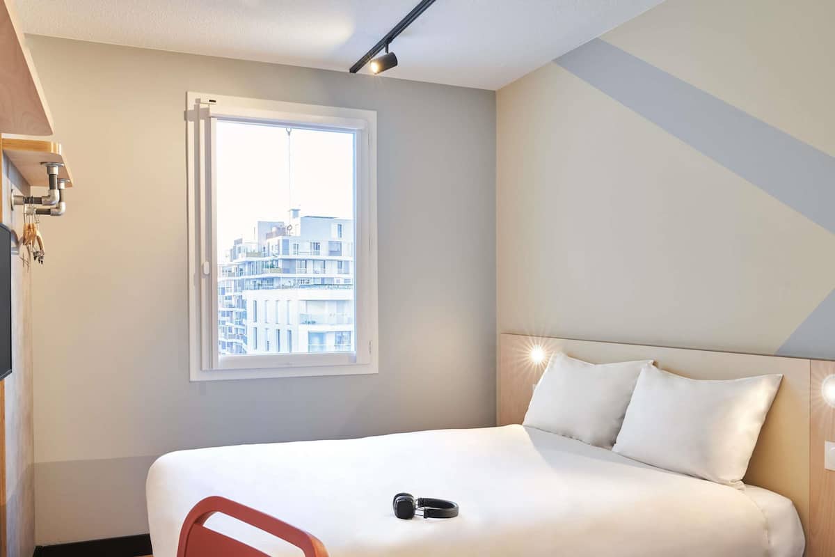 Une chambre d’hôtel avec un lit, une fenêtre donnant sur des immeubles, un fauteuil rouge et un bureau avec une lampe.