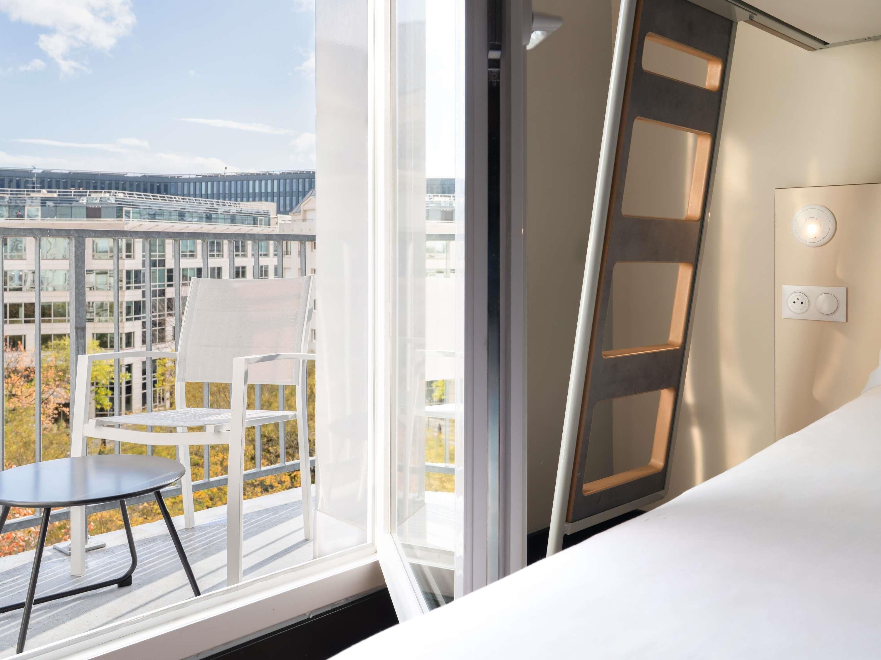 A modern hotel room with a balcony, a white chair, a small table, and a view of a cityscape.