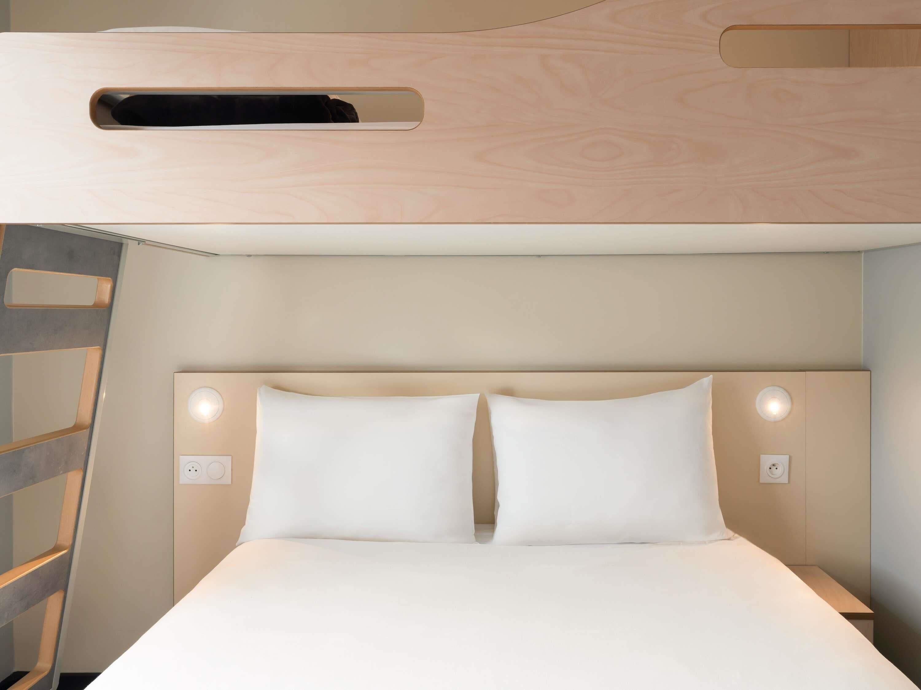 A bed with white bedding, a headboard with two wall-mounted lights, and a wooden ceiling with a ventilation panel.