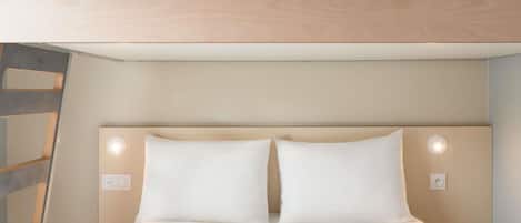 Una cama con ropa blanca, un cabecero con dos luces fijadas a la pared, y un techo de madera con un panel de ventilación.