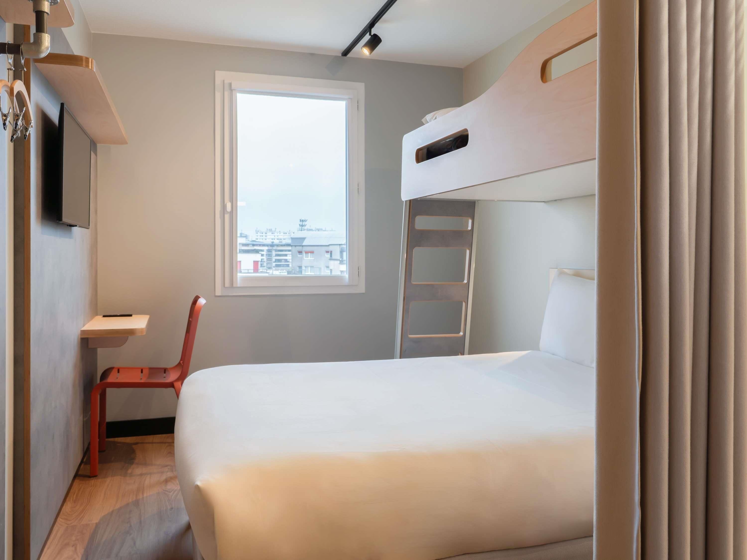 A compact hotel room with a bunk bed, a desk, a chair, a window, and a TV mounted on the wall.