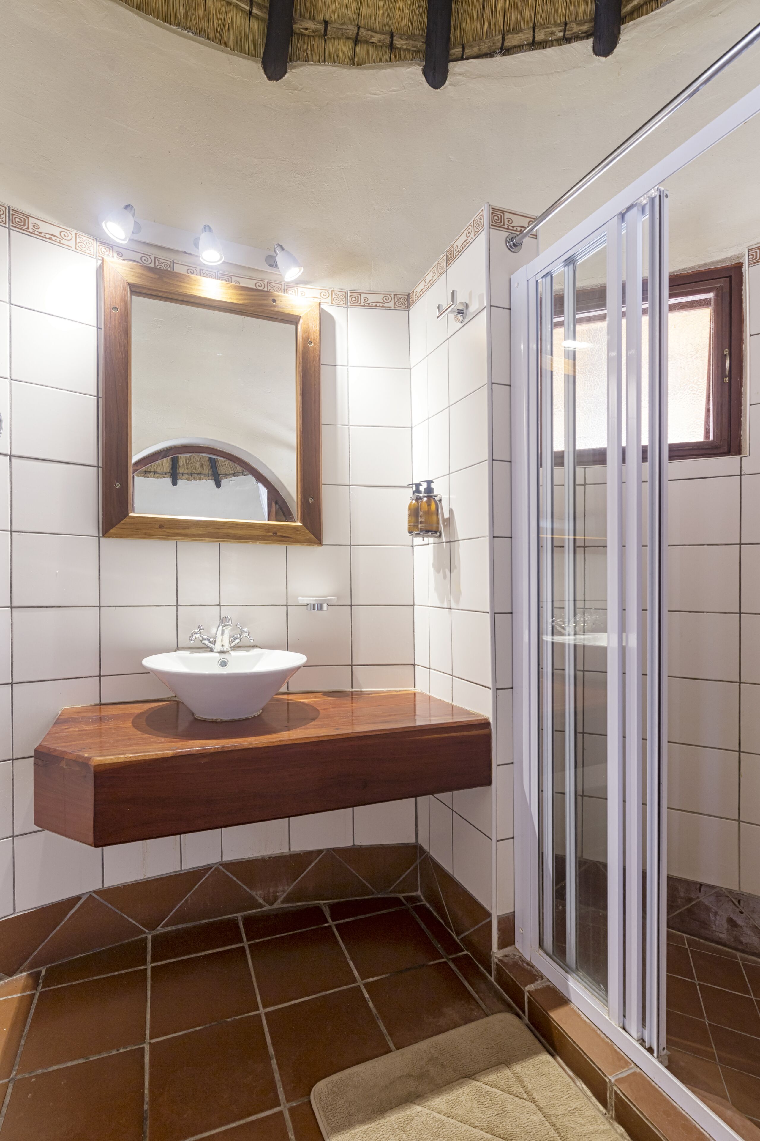 Un baño con tocador de madera, un lavabo blanco, un espejo con luces, ducha con puerta de vidrio y paredes con azulejos.