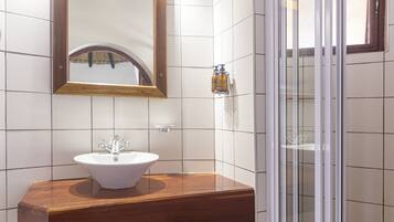 Un baño con tocador de madera, un lavabo blanco, un espejo con luces, ducha con puerta de vidrio y paredes con azulejos.