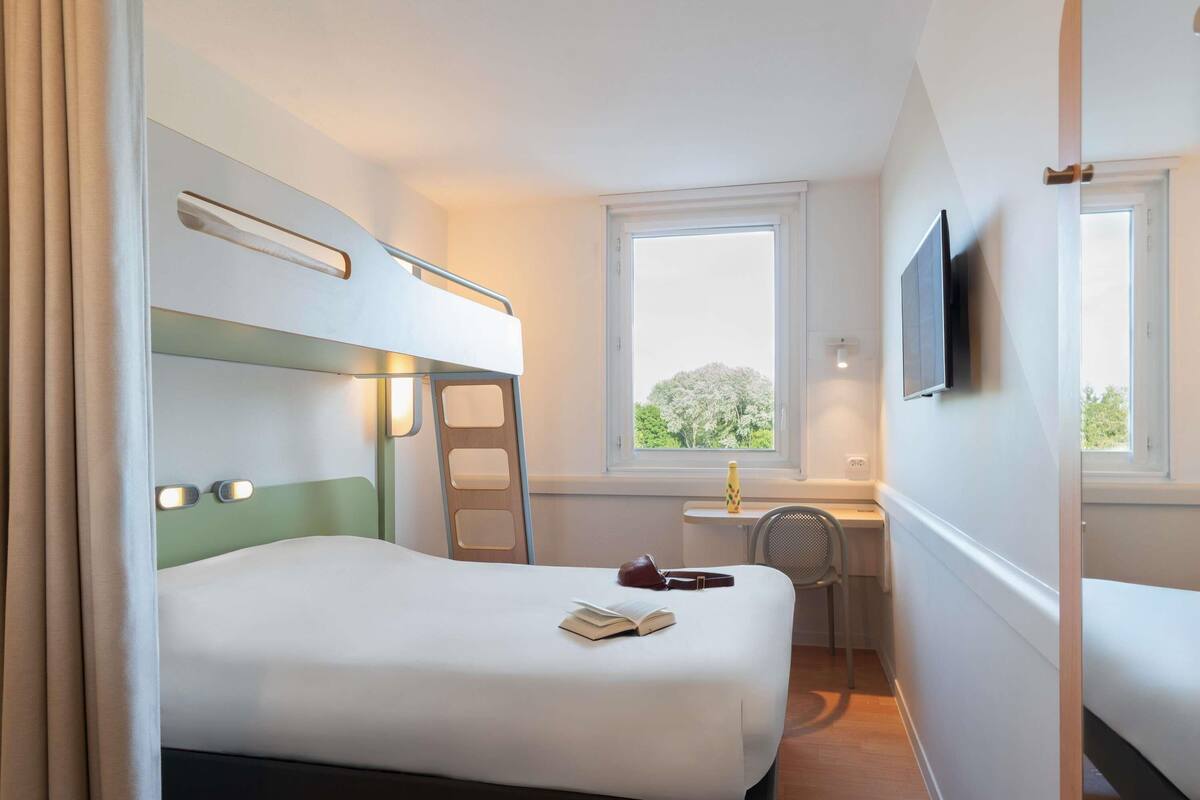 A hotel room with a bunk bed, a desk with a chair, a window with a view, and a TV mounted on the wall.