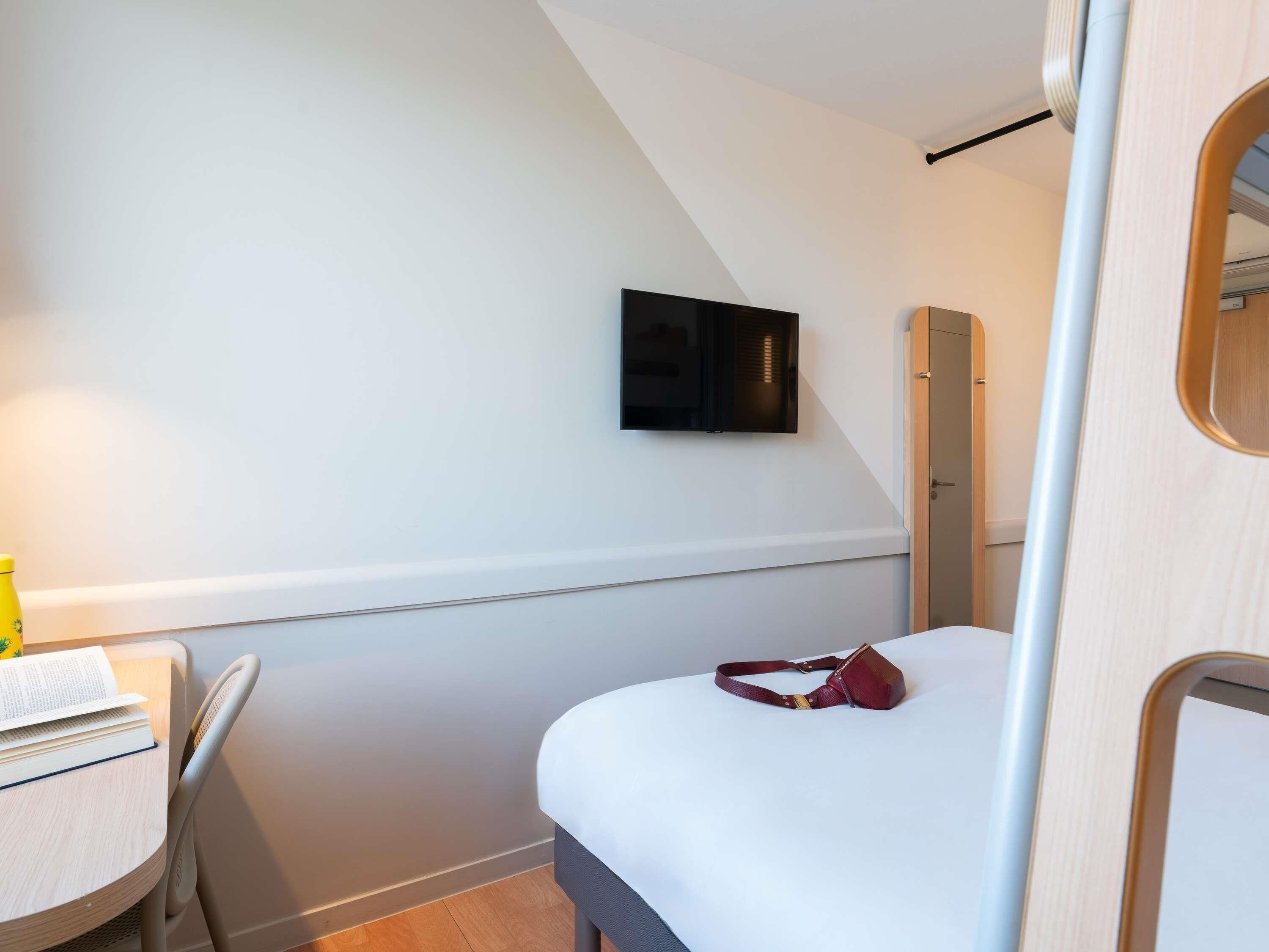 Habitació petita i moderna amb un llit, un escriptori amb cadira, una televisió penjada a la paret i una porta amb marc de fusta.
