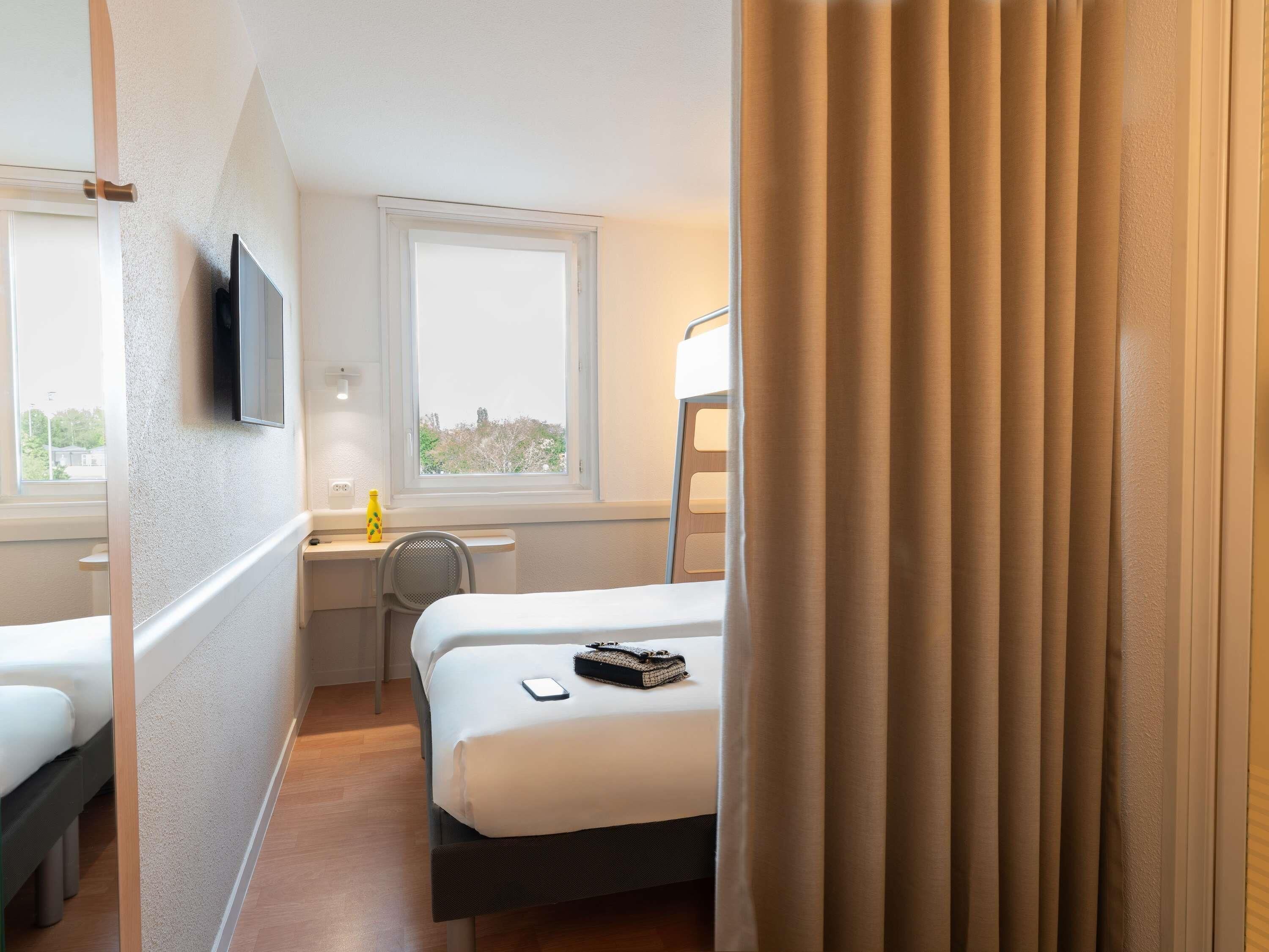 Une chambre d’hôtel avec deux lits, un bureau, une chaise, une télévision et une fenêtre avec des rideaux.