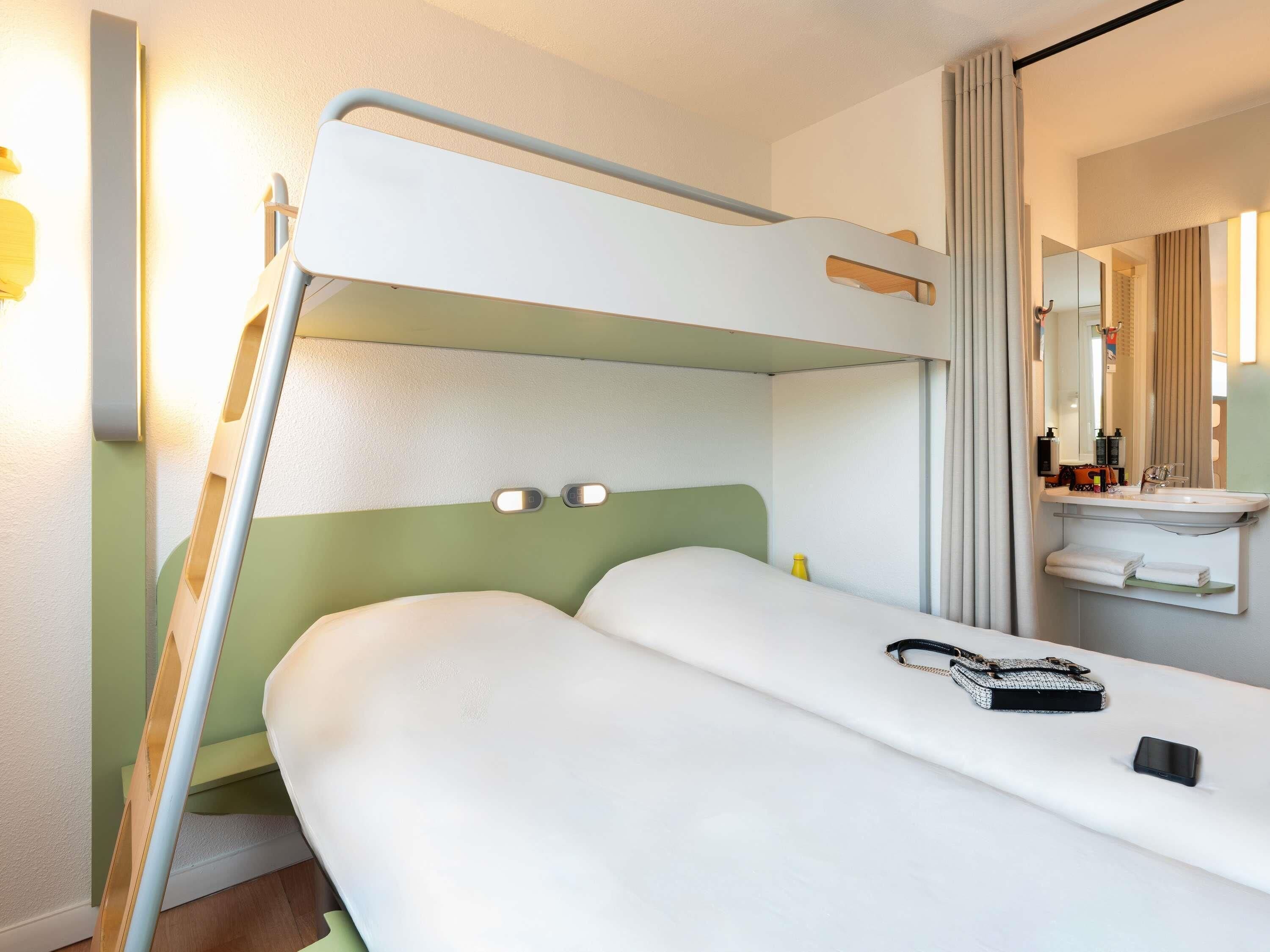 Een stapelbed met een groen hoofdeinde, een nachtkastje met een boek en een smartphone, en een badkamer met een wastafel en spiegel.