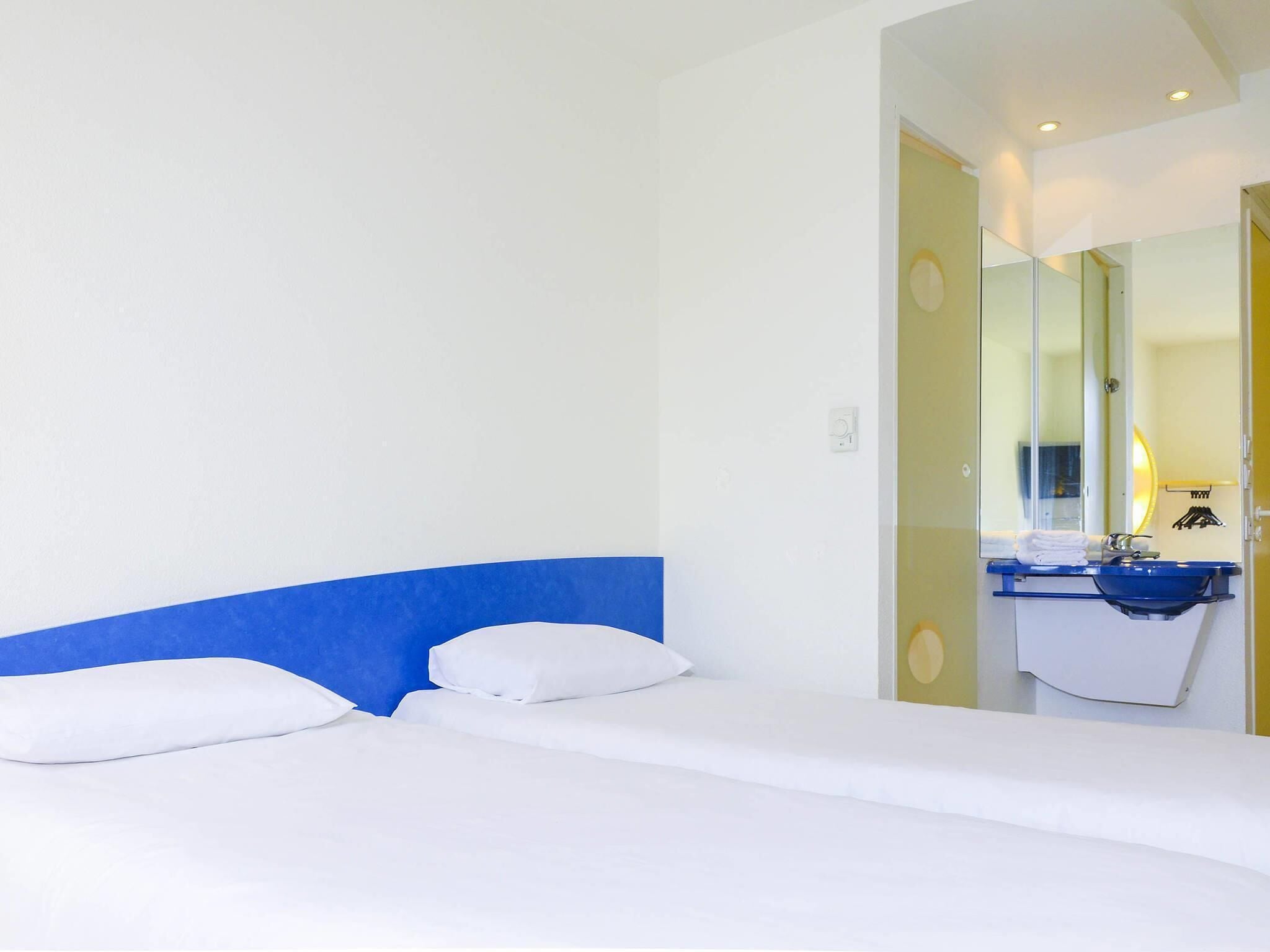 Ein Hotelzimmer mit Bett, blauem Kopfteil, Badezimmer mit Waschbecken und Spiegel sowie einer Tür zu einem weiteren Raum.
