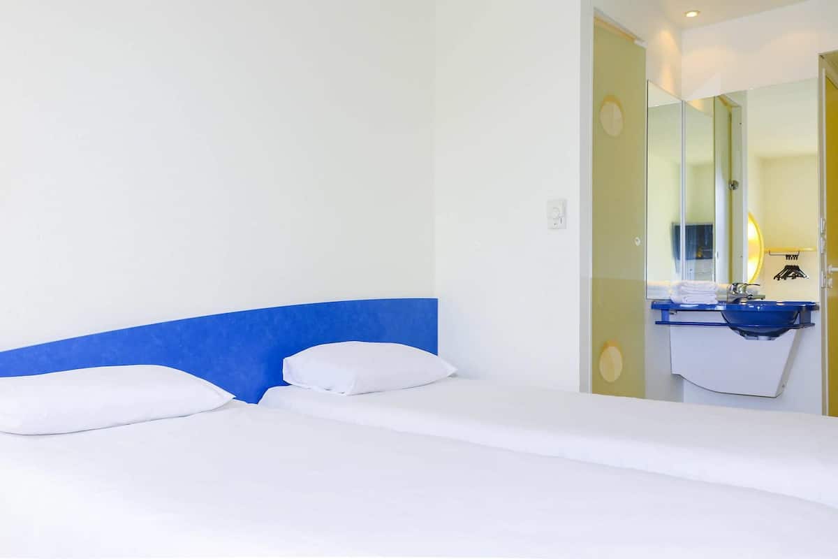 Ein Hotelzimmer mit Bett, blauem Kopfteil, Badezimmer mit Waschbecken und Spiegel sowie einer Tür zu einem weiteren Raum.