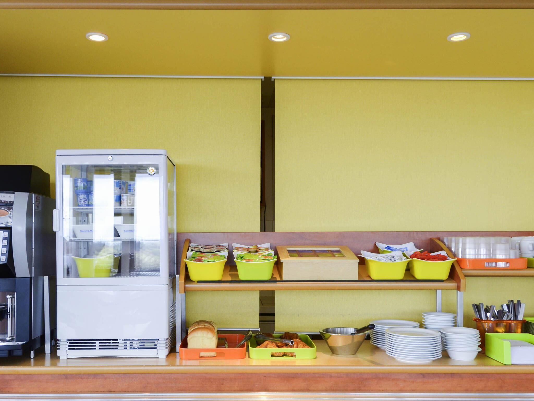 A breakfast buffet with a refrigerator, trays of food, and stacked plates.
