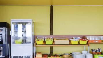 A breakfast buffet with a refrigerator, trays of food, and stacked plates.