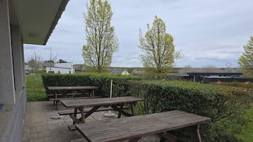 An outdoor patio with picnic tables, a hedge, and a building in the background.