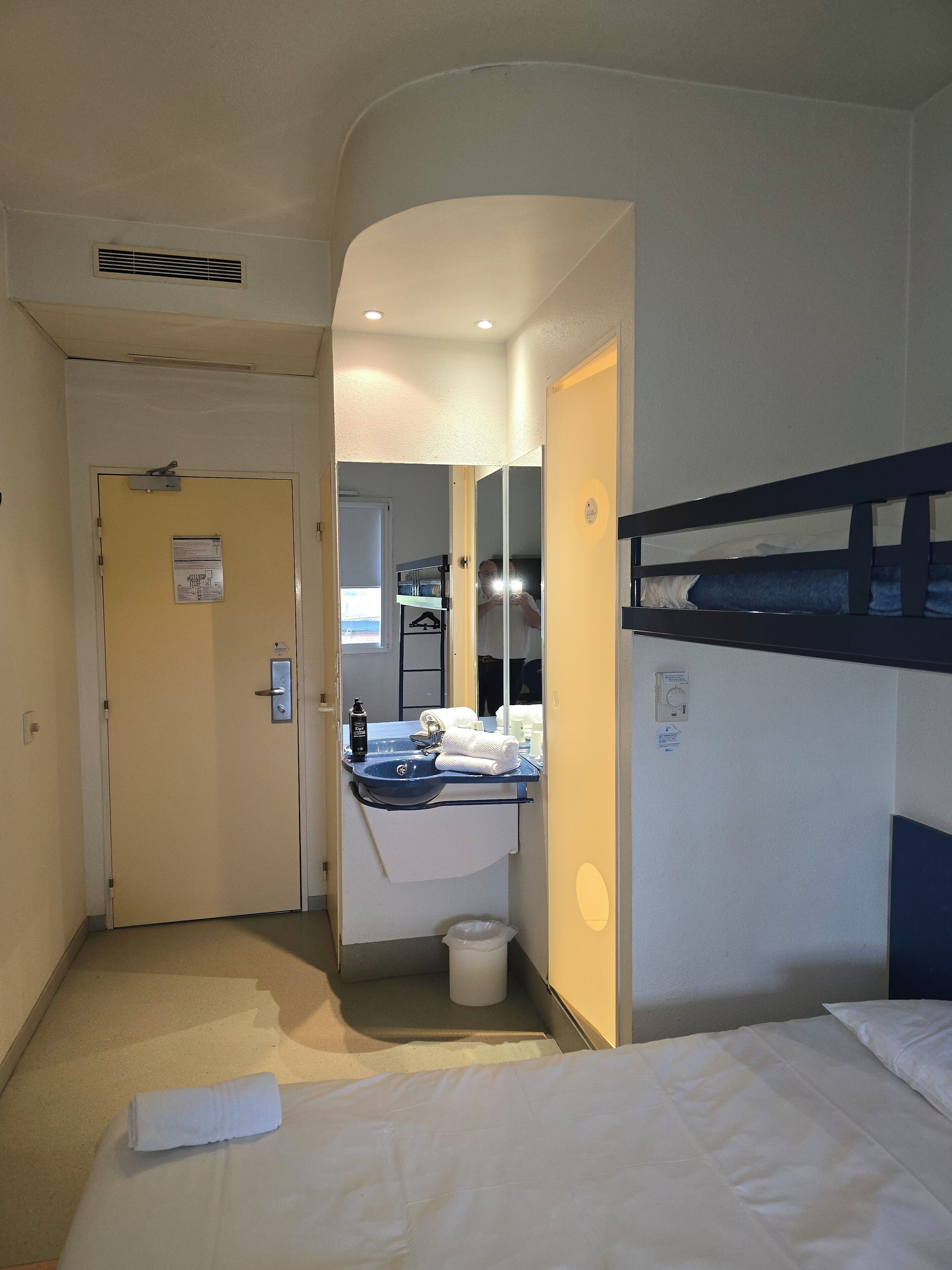 Quarto de hotel com uma cama beliche, banheiro com pia e espelho, uma porta com placa e uma pequena lixeira.