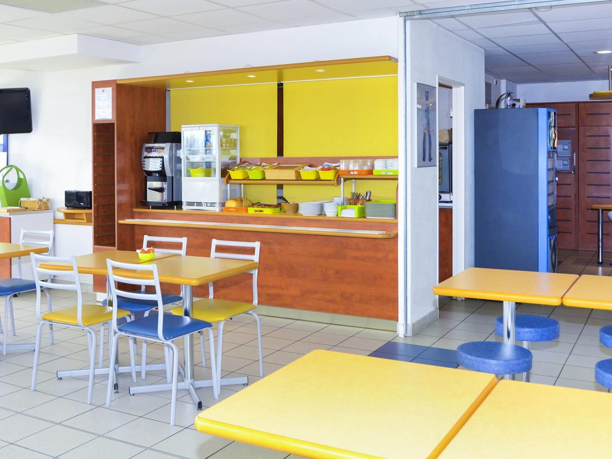 Une cafétéria avec des tables et des chaises jaunes et bleues, un réfrigérateur et une machine à café.