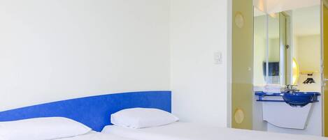 Habitación de hotel con cama, cabecera azul, baño con lavabo y espejo, y una puerta que da a otra habitación.