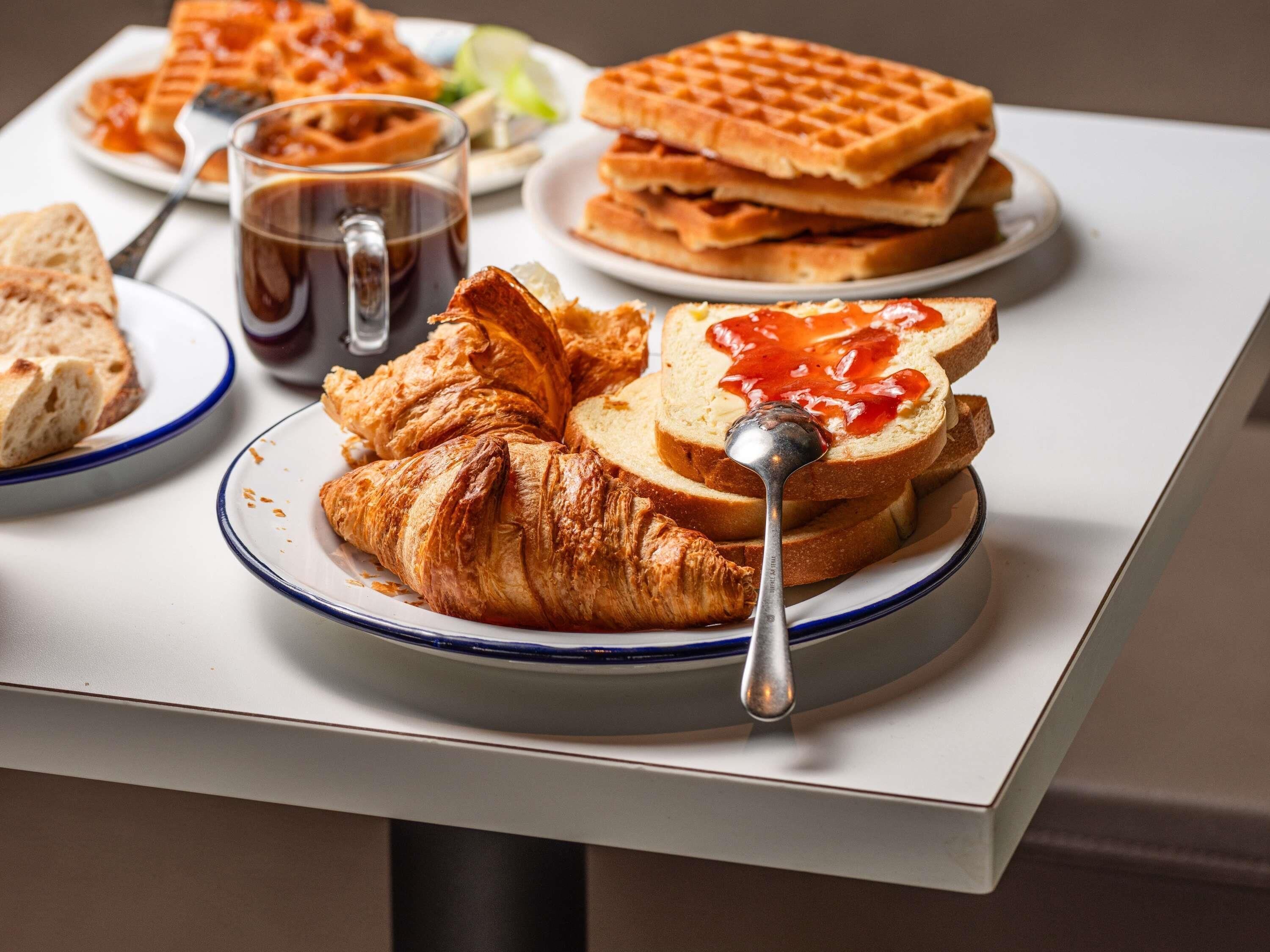 Un desayuno con croissants, gofres, tostadas con mermelada y una taza de café sobre una mesa.