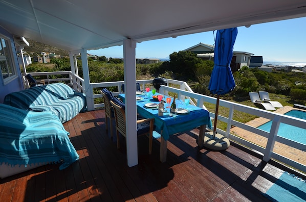 Un espacio exterior techado con zona de estar, mesa lista para comer, piscina y vistas del paisaje circundante.