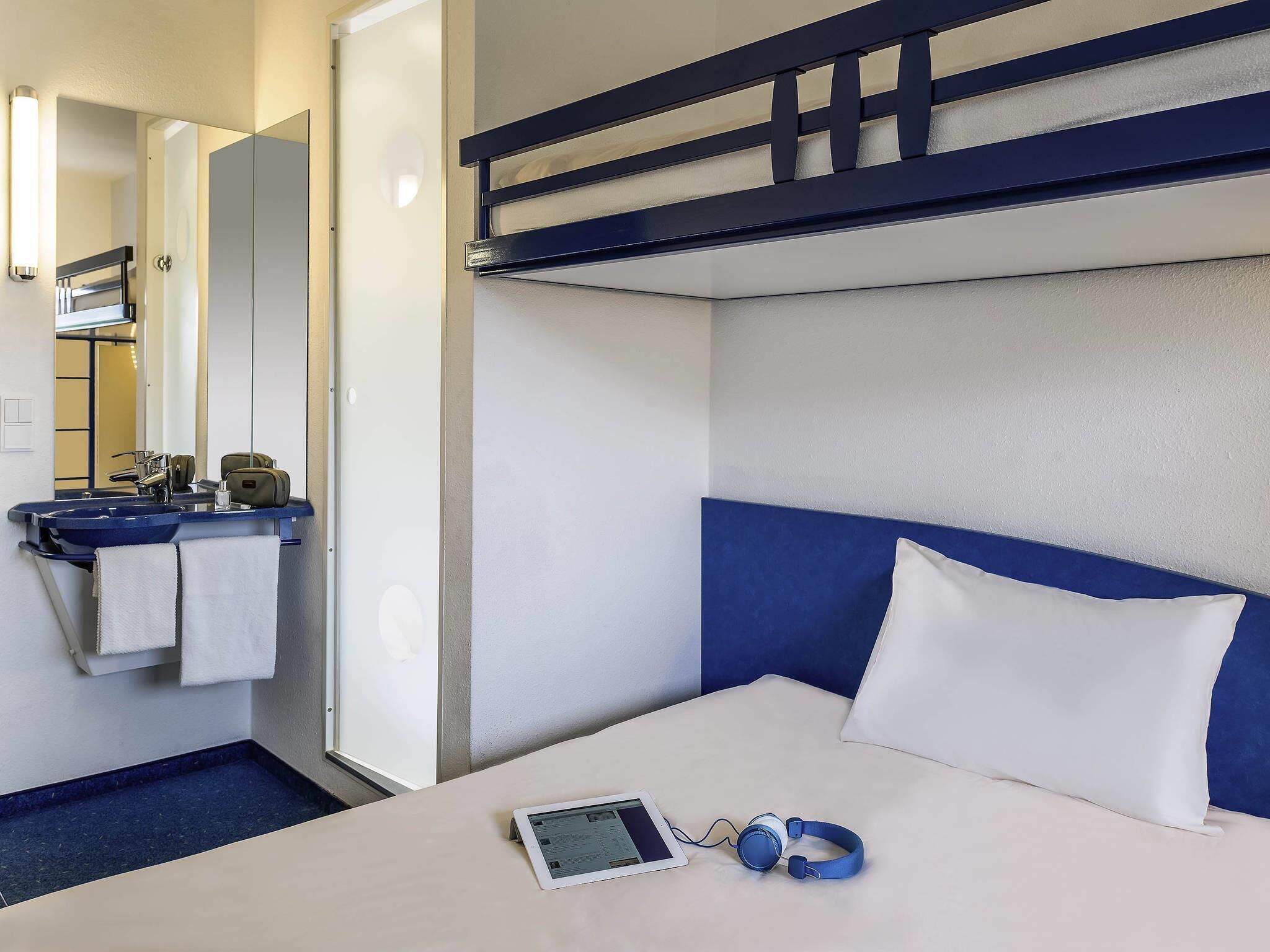 A hotel room with a bunk bed, a bedside table with a tablet and headphones, a sink with a towel, and a mirror.