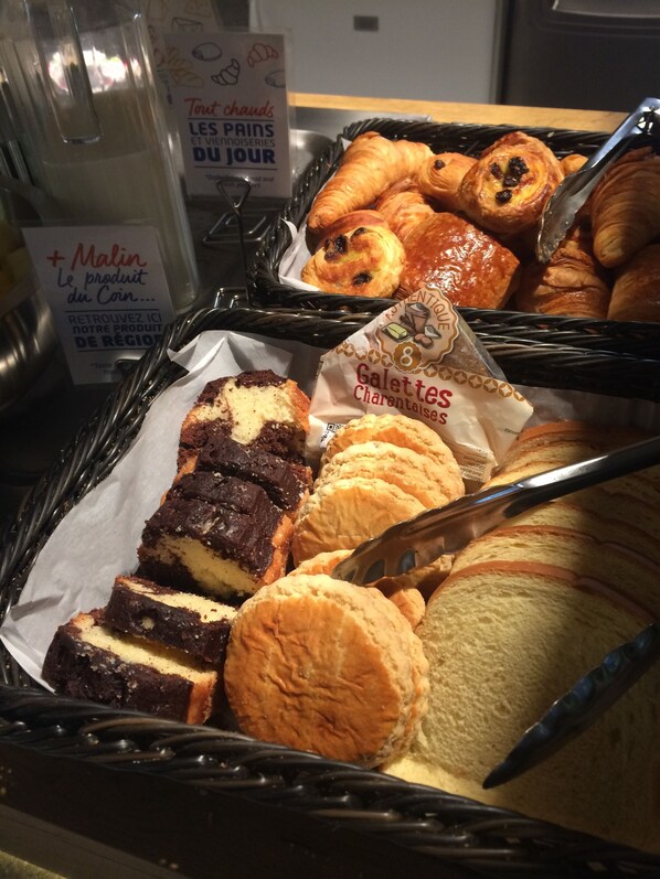 A display of assorted pastries and baked goods, including croissants, muffins, and a chocolate cake, arranged in a basket. - ibis budget Rochefort (Rochefort)