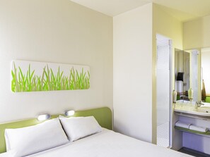 Habitación de hotel con cama, luz empotrada en la pared, espejo y baño con lavamanos y perchero para toallas.
