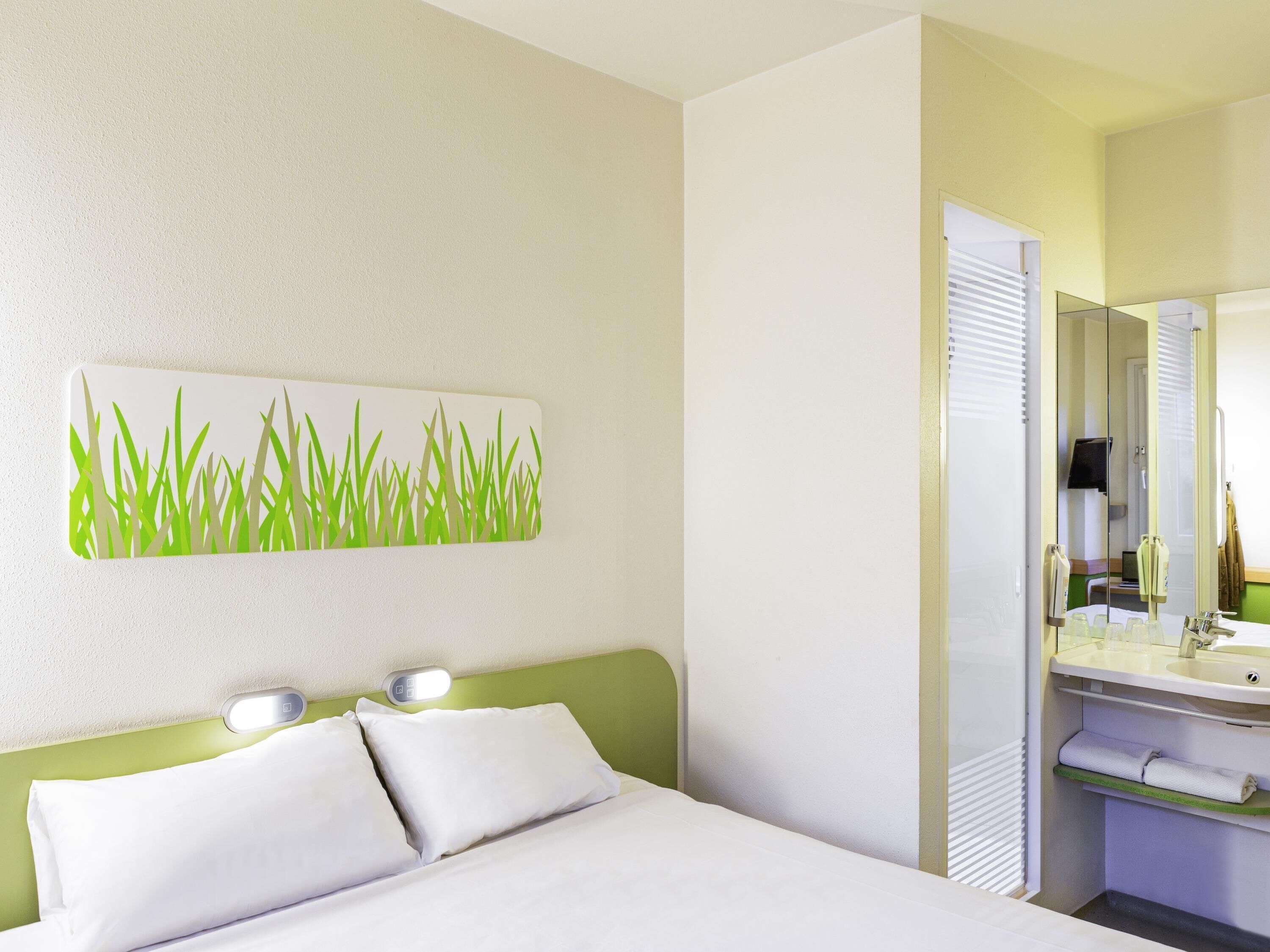 A hotel room with a bed, a wall-mounted light, a mirror, and a bathroom with a sink and towel rack.