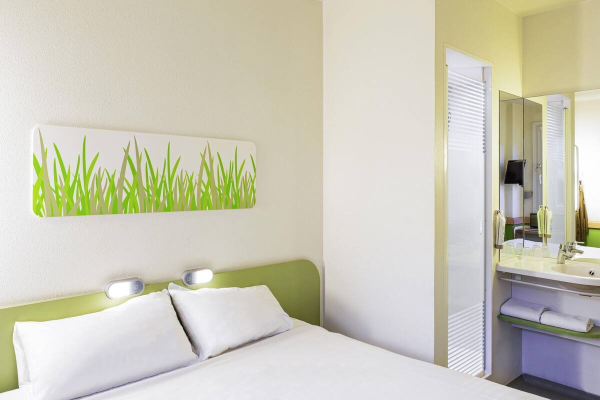 A hotel room with a bed, a wall-mounted light, a mirror, and a bathroom with a sink and towel rack.