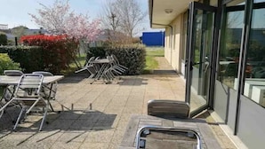 An outdoor patio area with tables and chairs, a small garden with flowering plants, and a building entrance with glass doors. - ibis budget Bergerac (Bergerac)